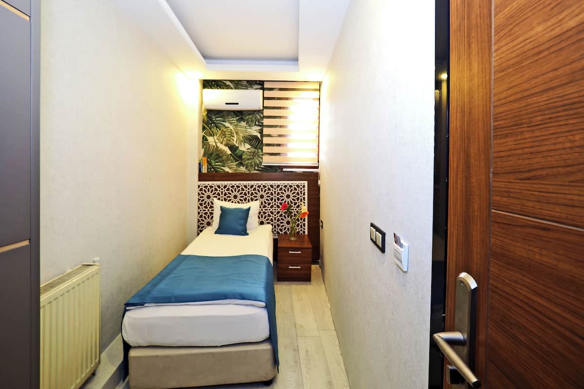 A narrow hotel room with a bed, a nightstand, a wall-mounted air conditioner, and a wooden door.