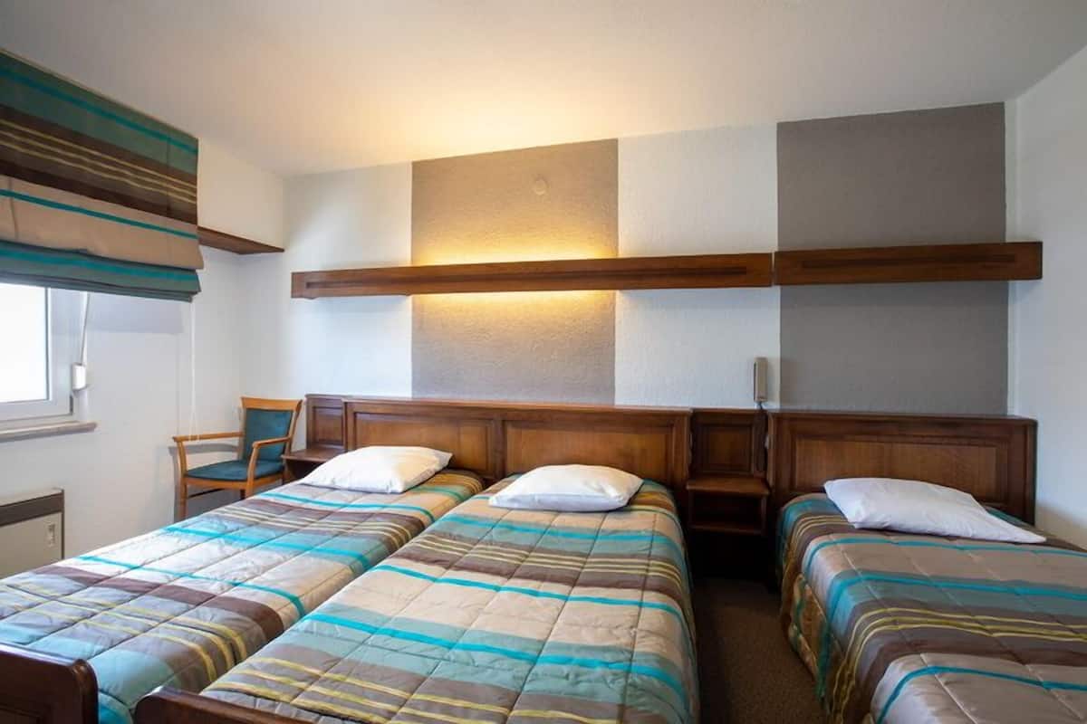 Una habitación de hotel con dos camas individuales, cabeceros de madera, una lámpara fijada a la pared y una ventana con cortinas.