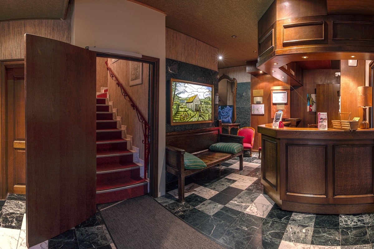 Un vestíbulo de hotel con una escalera de madera, una recepción, un mostrador de registro, una zona de estar con un banco y sillas, y un cuadro grande en la pared.