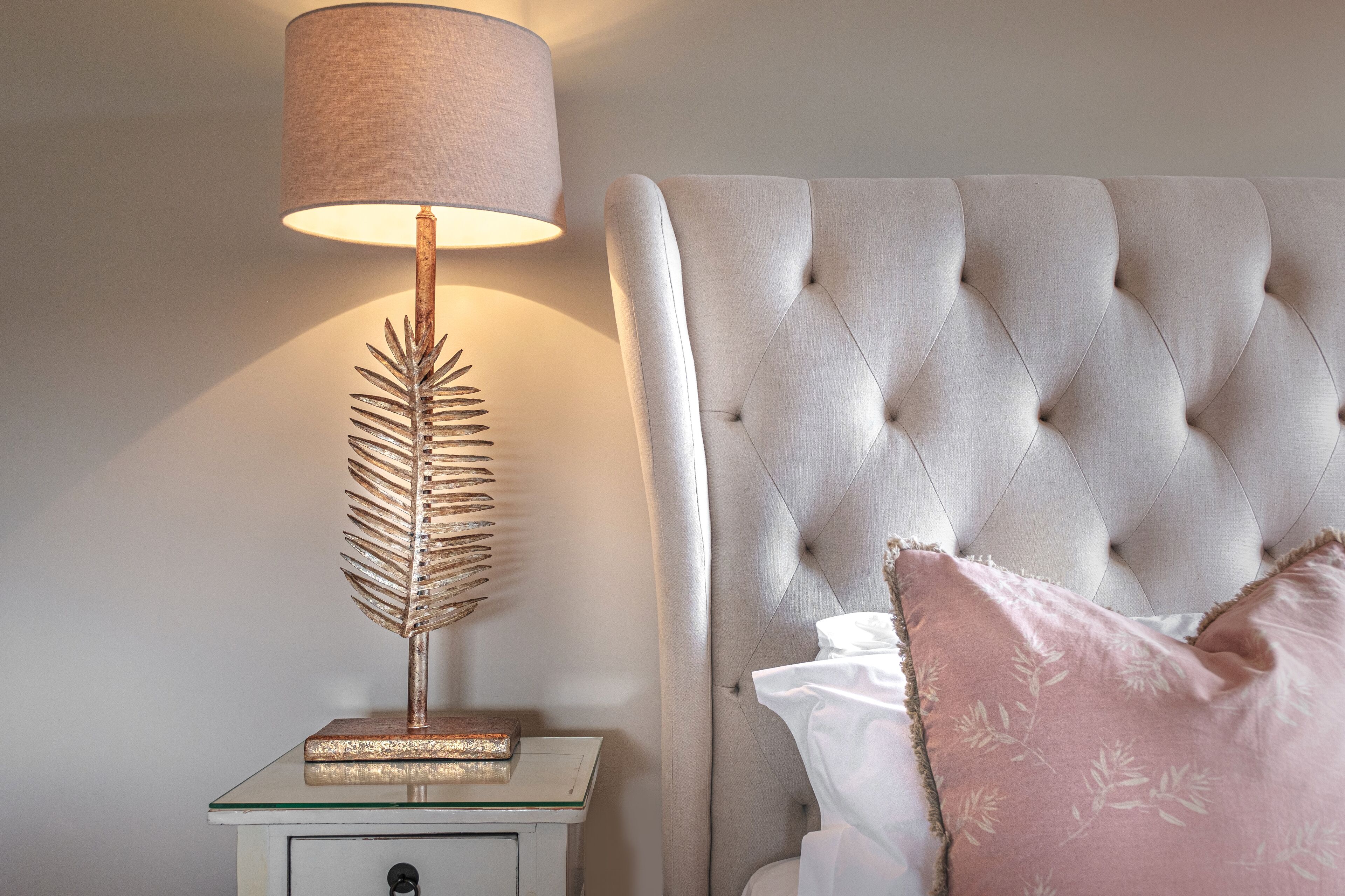 A bedroom with a tufted headboard, a bedside lamp with a leaf-shaped base, and a white nightstand.