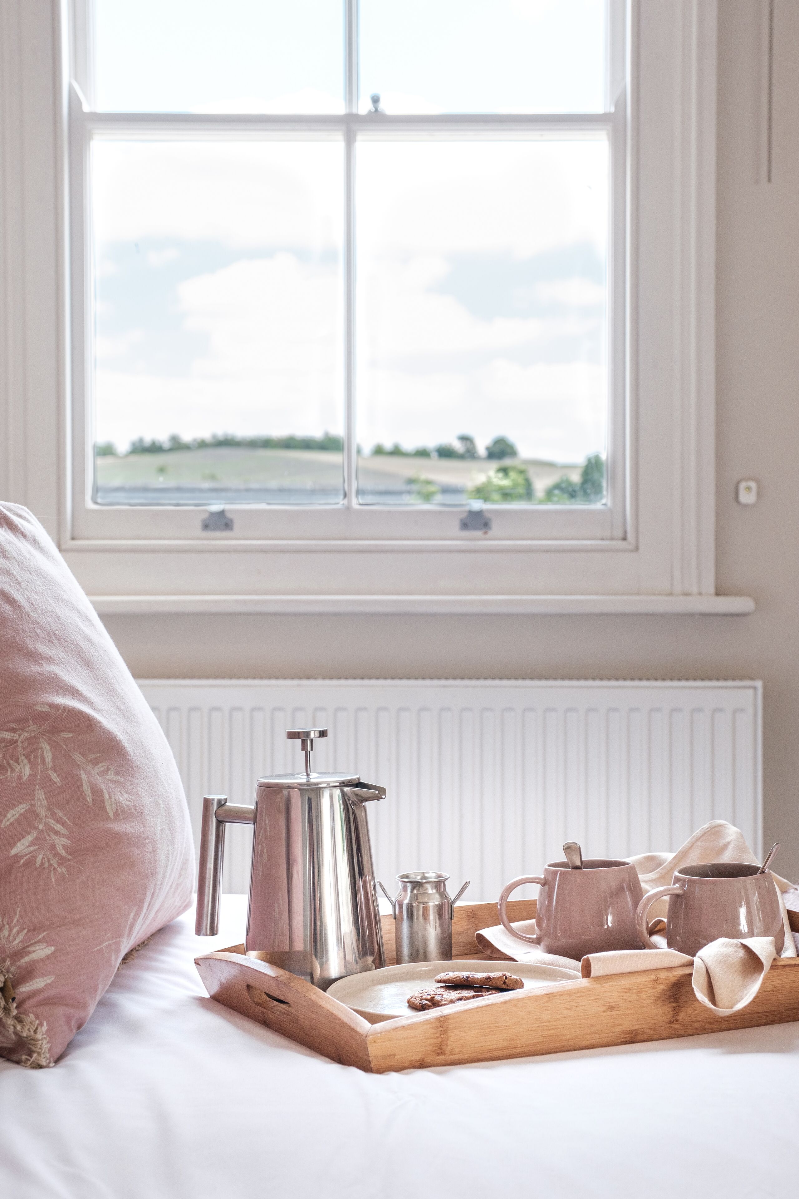 Una bandeja de desayuno bien dispuesta con café y pasteles en una cama cerca de una ventana con vistas a un campo.