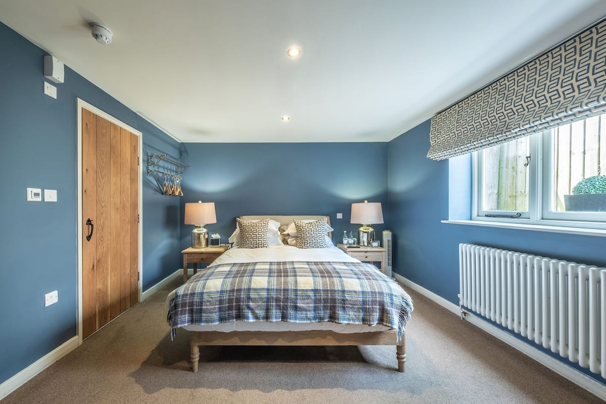 A bedroom with a bed, two bedside tables, a window with blinds, a radiator, and a door.