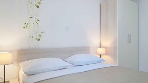 A neatly made bed with white linens, a bedside lamp, a wall-mounted plant, and a wardrobe. - Holiday apartment mit Pool (Baška)