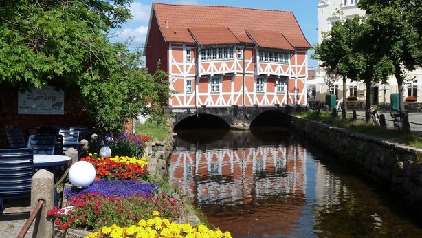 Un edificio tradicional con entramado de madera y techo rojo, un puente de piedra y un canal con flores coloridas al frente.