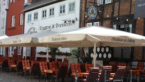 Un café al aire libre con mesas y sillas, un letrero que dice 'Brauhaus am Löffberg' y arquitectura europea tradicional al fondo.