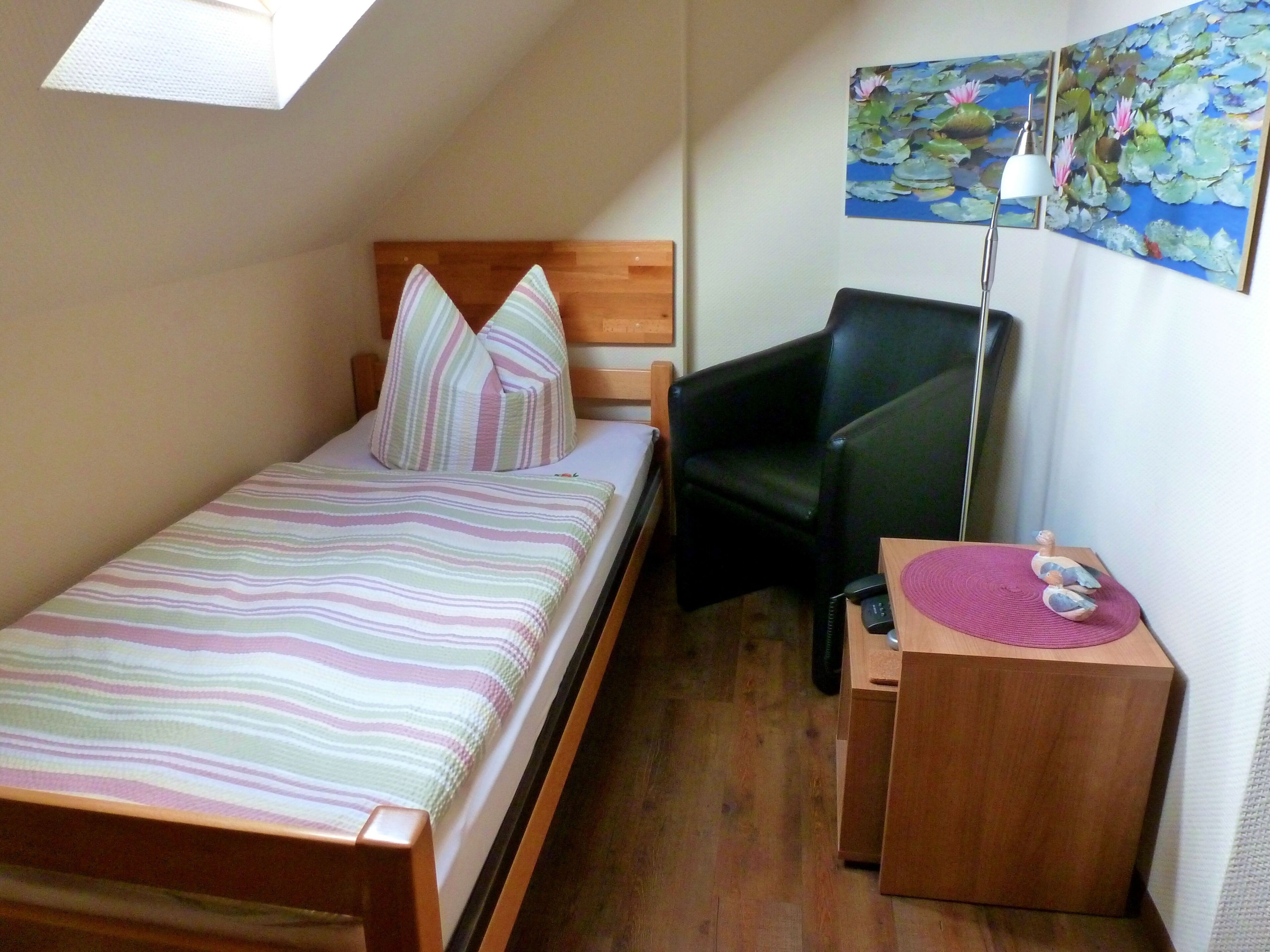Une petite chambre avec un lit simple, une tête de lit en bois, une couvre-lit à rayures, une chaise noire, un bureau avec une lampe et un tableau représentant des nénuphars accroché au mur.