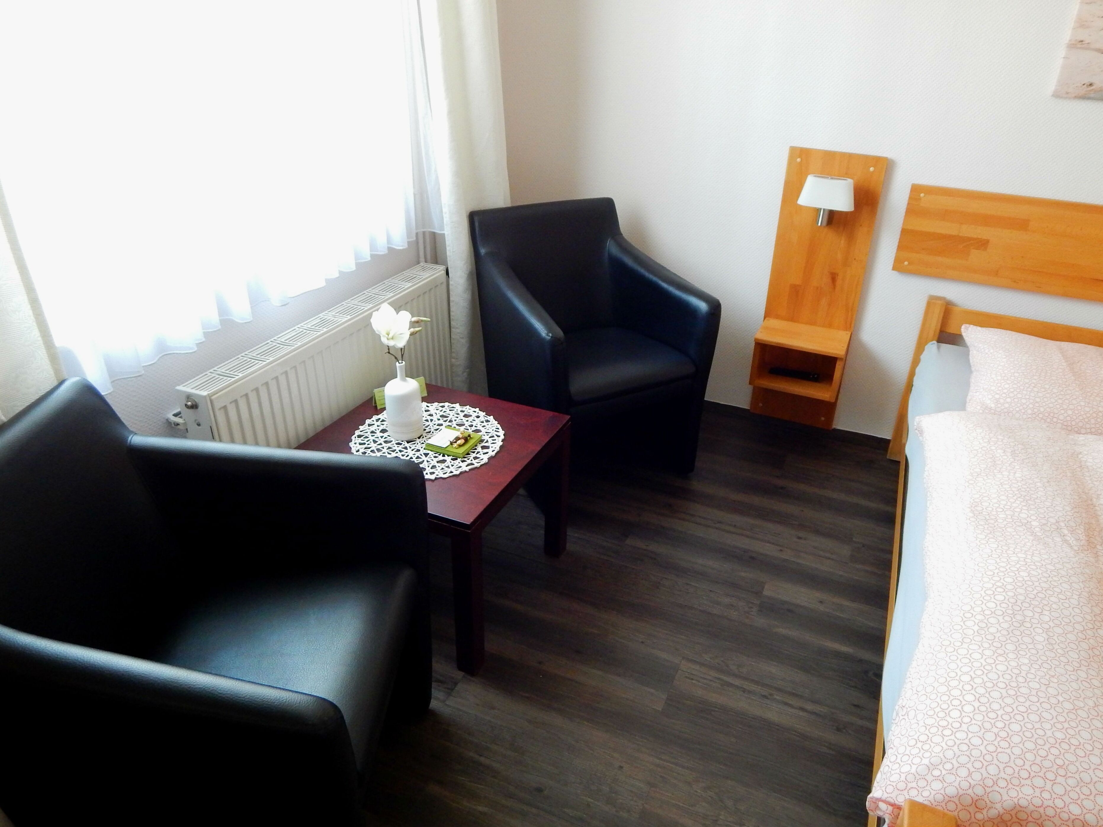A hotel room with a bed, a desk with a chair, a nightstand with a lamp, and a window with curtains.