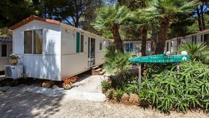 Uma casa móvel com um pequeno alpendre, rodeada por árvores e plantas.