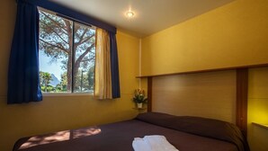 Uma cama de solteiro com colchão marrom e toalha branca, uma janela com cortinas azuis e uma pequena planta sobre uma mesinha de cabeceira.