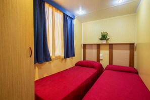 A room with two single beds, a wooden wardrobe, a window with blue curtains, and a small table with a plant. - Camping Village El-Bahira (San Vito Lo Capo)