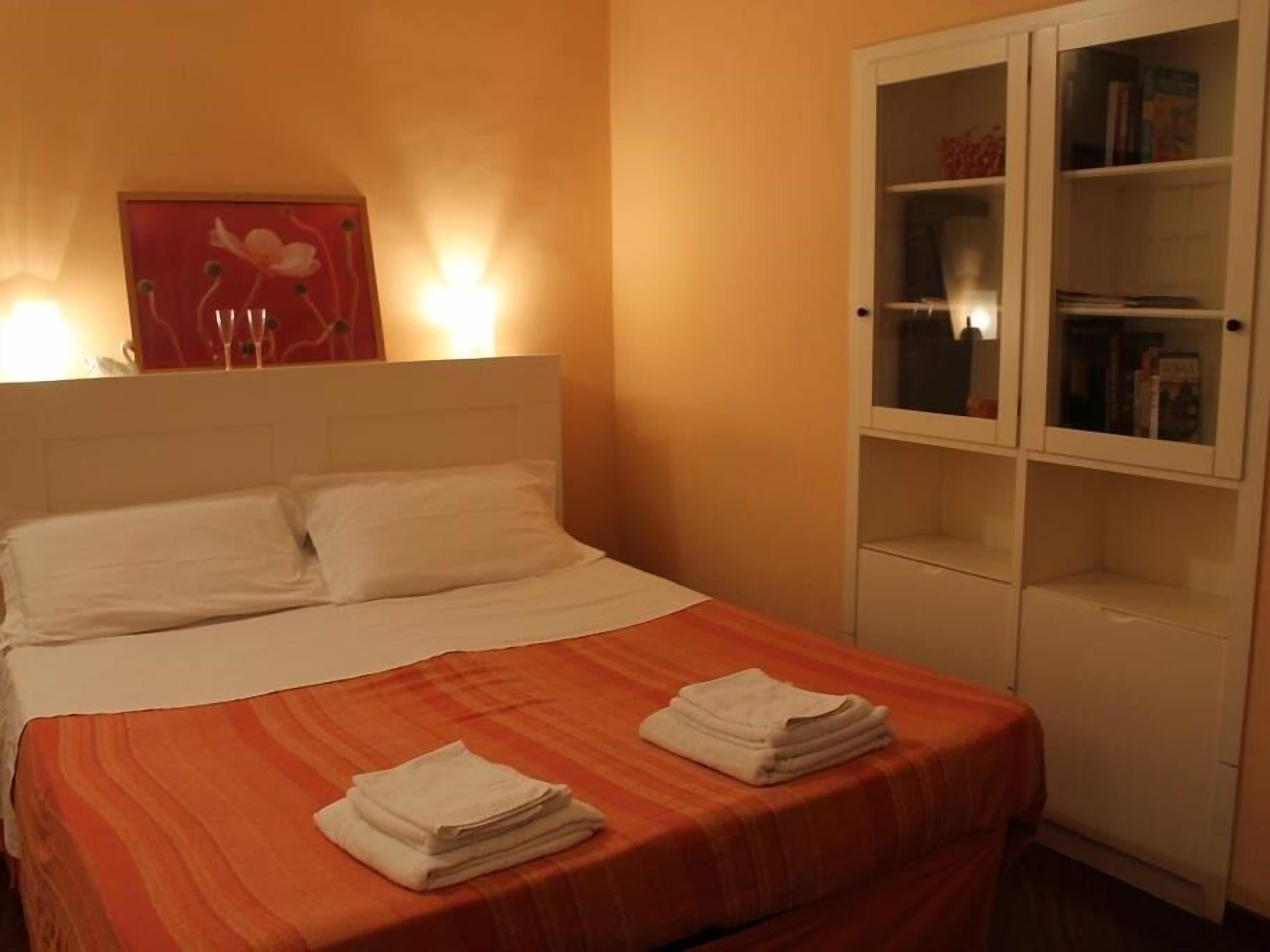 A neatly made bed with white pillows and an orange bedspread, a white wardrobe with glass doors, and a wall-mounted light fixture.