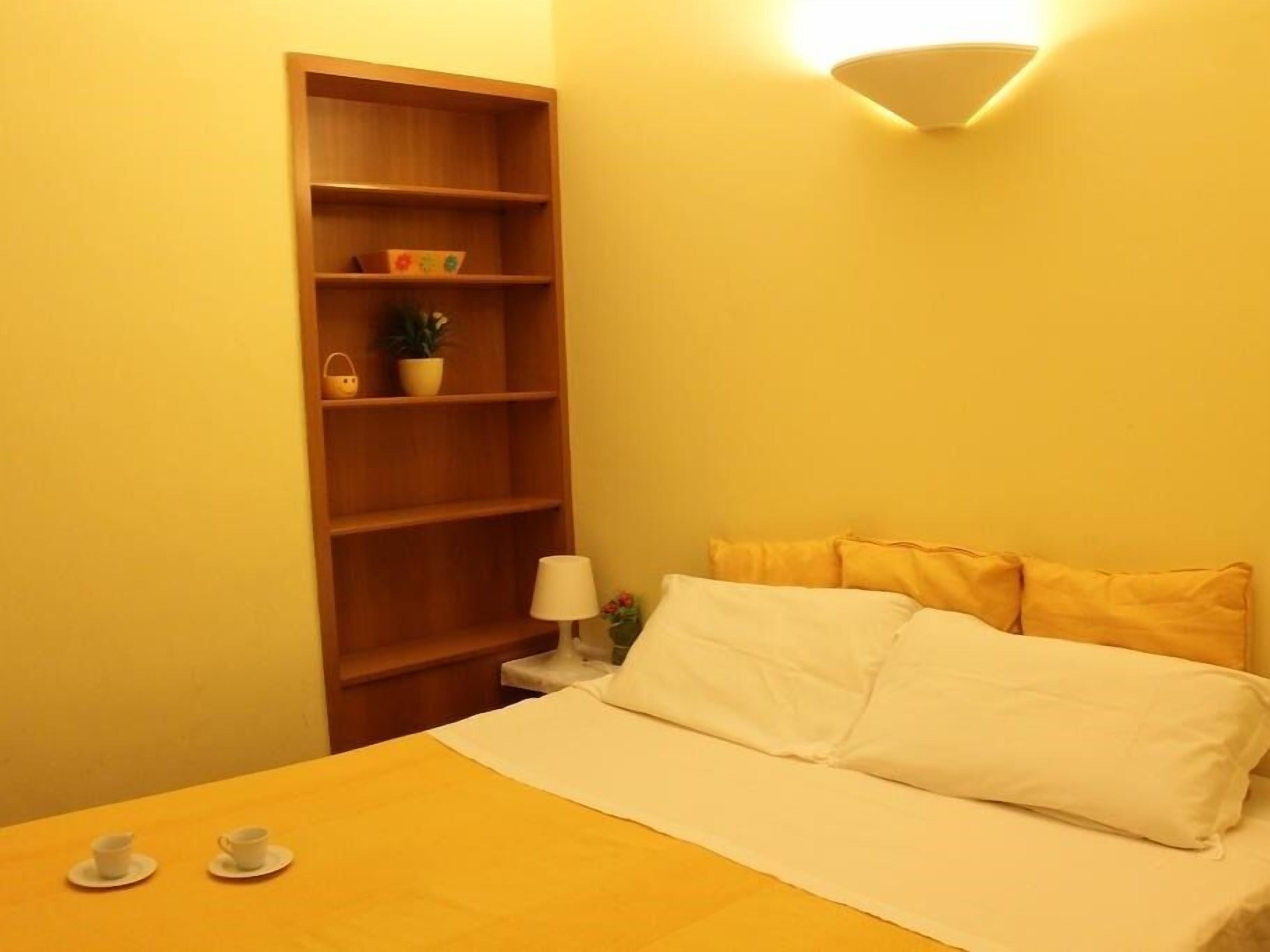 A bedroom with a bed, a nightstand, a lamp, a shelf with decorative items, and two cups on the bed.
