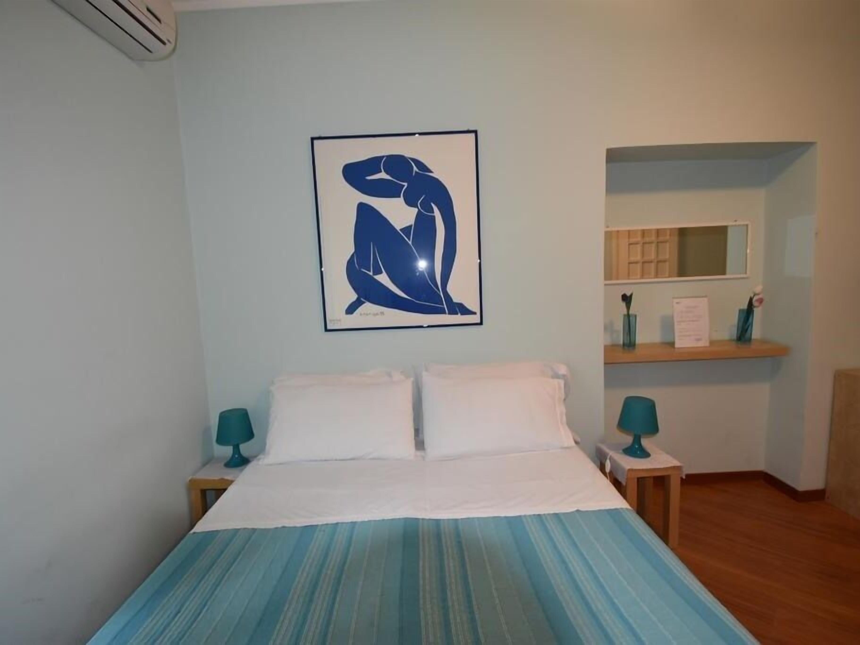 A bedroom with a bed, two bedside tables, a nightstand, a wall-mounted air conditioner, a framed abstract artwork, a mirror, and a shelf with a vase and a sign.