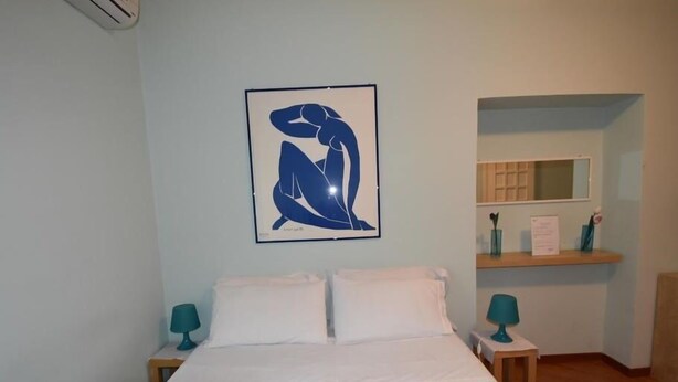 A bedroom with a bed, two bedside tables, a nightstand, a wall-mounted air conditioner, a framed abstract artwork, a mirror, and a shelf with a vase and a sign. - B&B A Casa Di Nancy (Rome)
