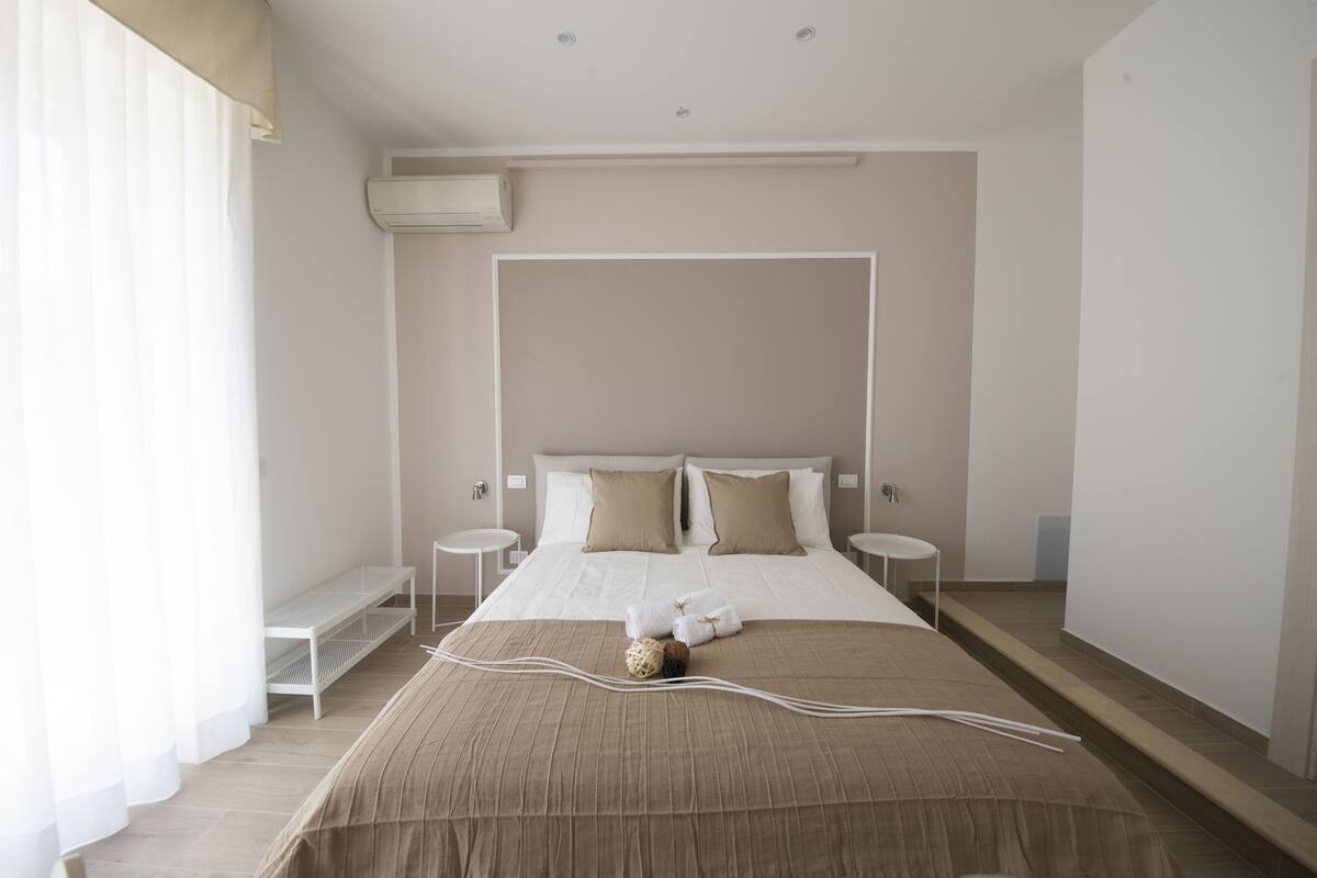 A modern hotel room with a large bed, two bedside tables, a bench, and an air conditioning unit.