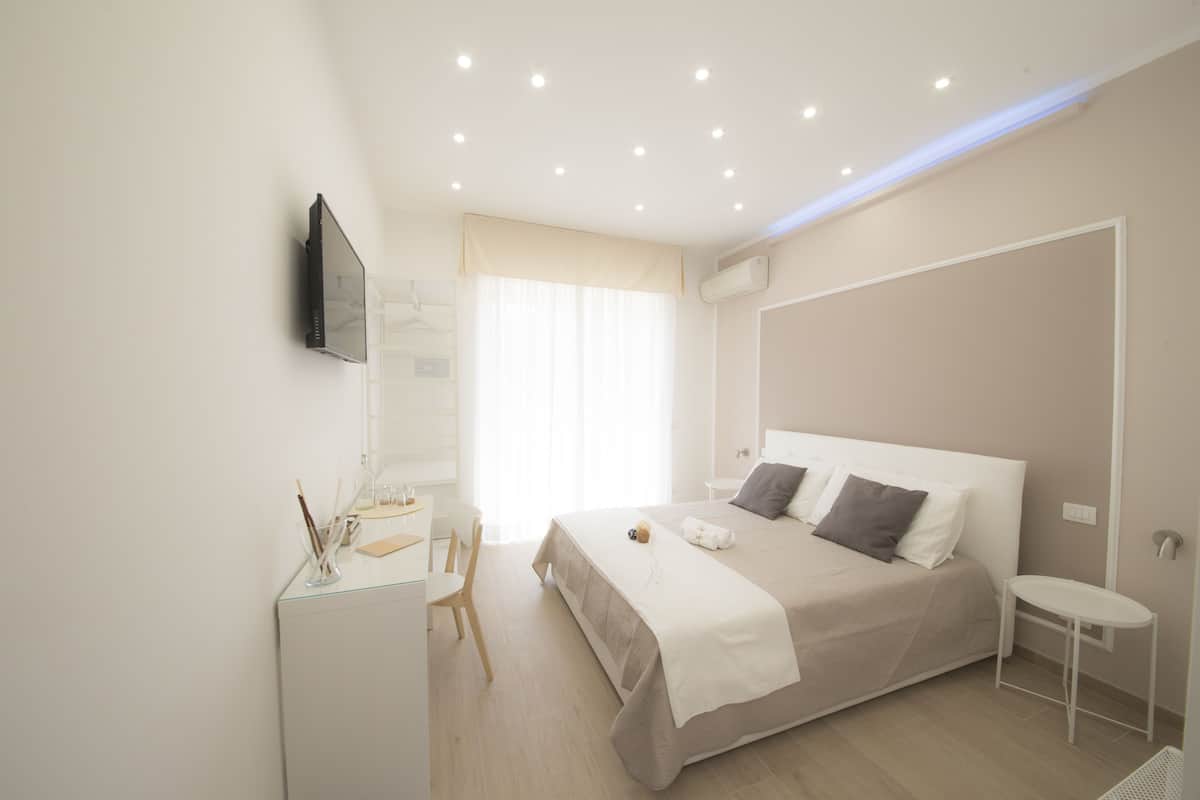 A modern hotel room with a large bed, a desk with a chair, a TV mounted on the wall, and a window with curtains.