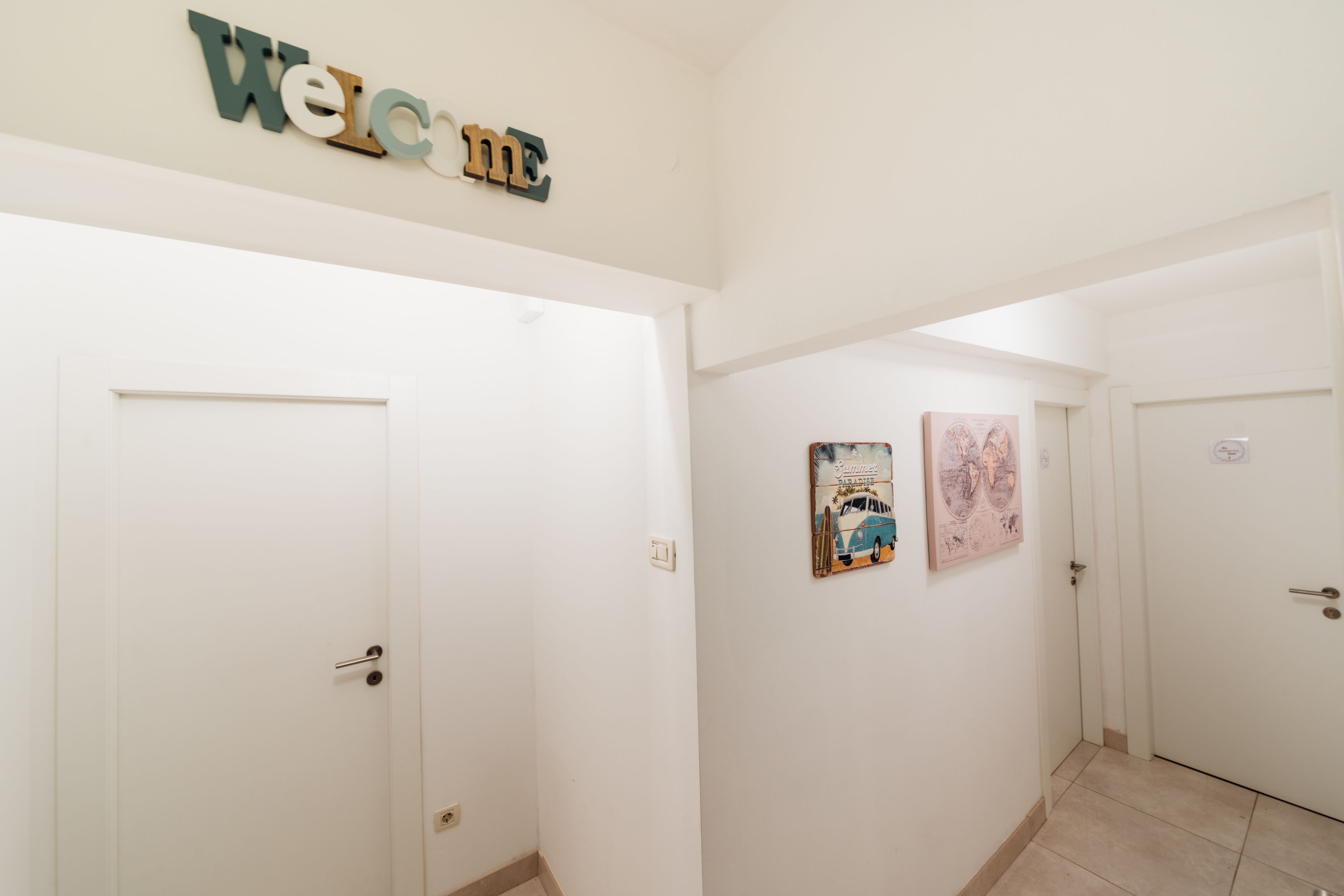 A hallway with white doors, a 'Welcome' sign, and two framed pictures on the wall.