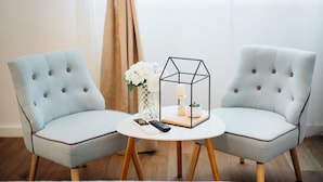 Two light blue upholstered chairs with wooden legs, a white round table, a vase with white flowers, and a geometric candle holder. - The Harmony Rooms Split (Split)