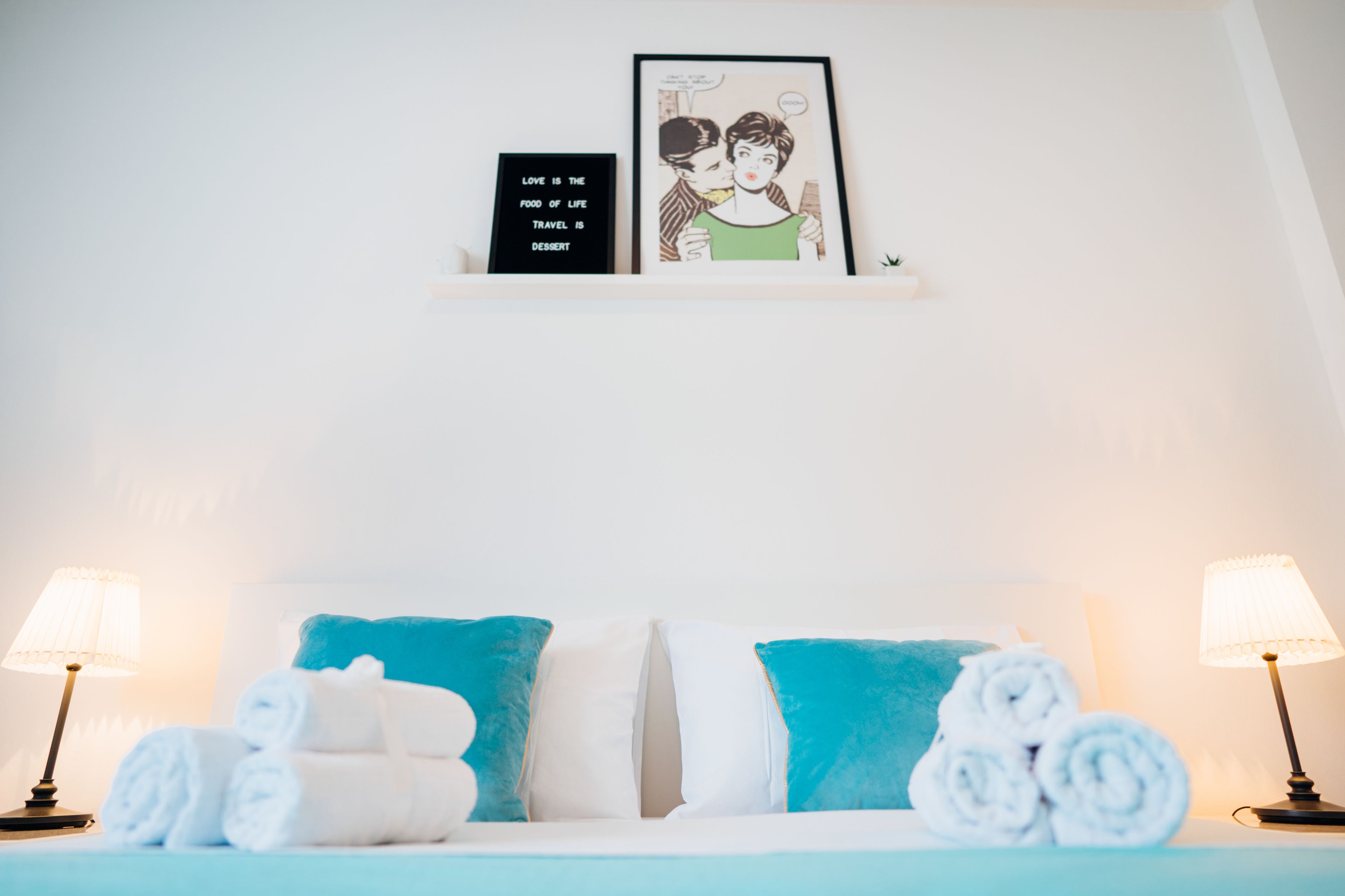 Een opgemaakt bed met blauw-witte beddengoed, twee opgerolde handdoeken, een ingelijst citaat en twee bedlampen.
