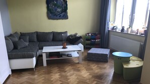A living room with a sofa, a coffee table, and a small table with a lamp. There is a window with curtains, a radiator, and a shelf with plants and toys. - Families, groups, cycling, biking, hiking, vacation with dog, short vacation, Kyllradweg, (Stadtkyll)