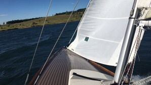 Cubierta de un velero con vela blanca, tablas de madera y una vista despejada del agua y la costa lejana.