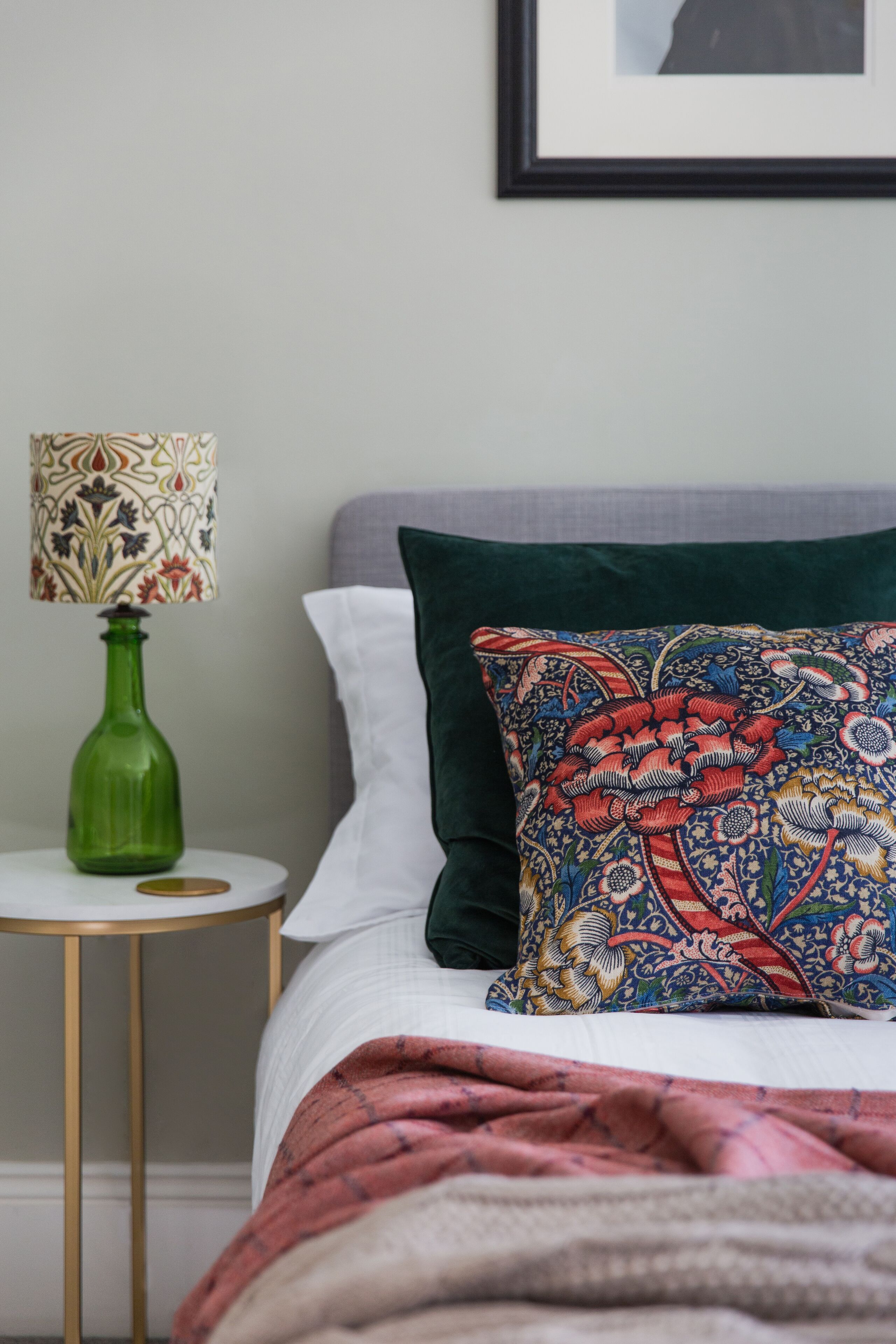 A neatly made bed with a patterned pillow, a green vase on a small round table, and a framed picture on the wall.