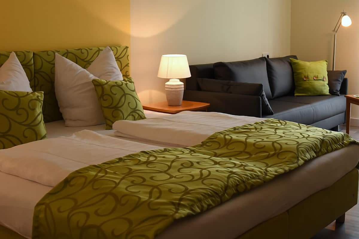 Una cama bien hecha con ropa de cama verde estampada, una lámpara de noche, un sofá oscuro con cojines verdes, y una lámpara sobre una mesita de madera.
