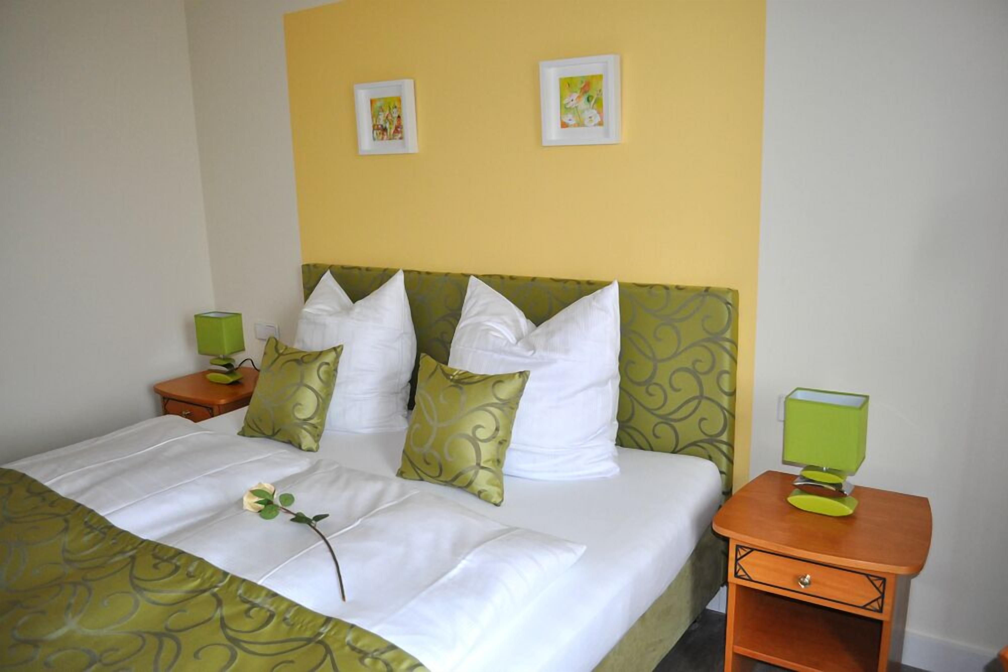 A neatly made bed with white and green bedding, two bedside tables with green lamps, and two framed pictures on the wall.