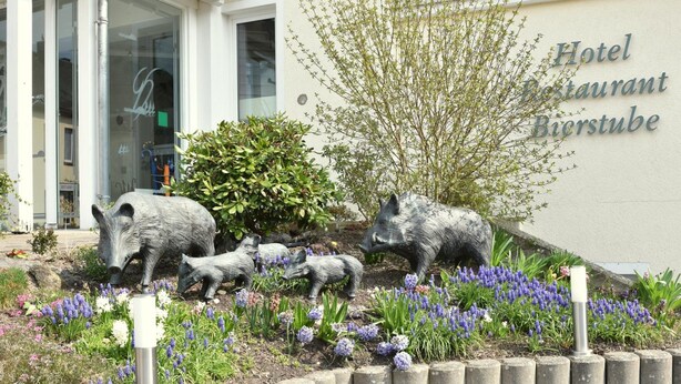 A building with a sign reading "Hotel Restaurant Bierstube" and a garden with pig sculptures and flowers. - Landhotel Gillenfelder Hof (Gillenfeld)