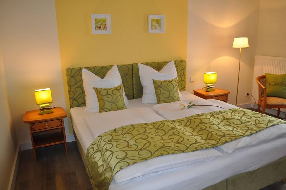 Una cama bien tendida con una colcha verde estampada, dos almohadas blancas y un cabecero verde a juego. Hay dos lámparas de noche, una a cada lado de la cama.
