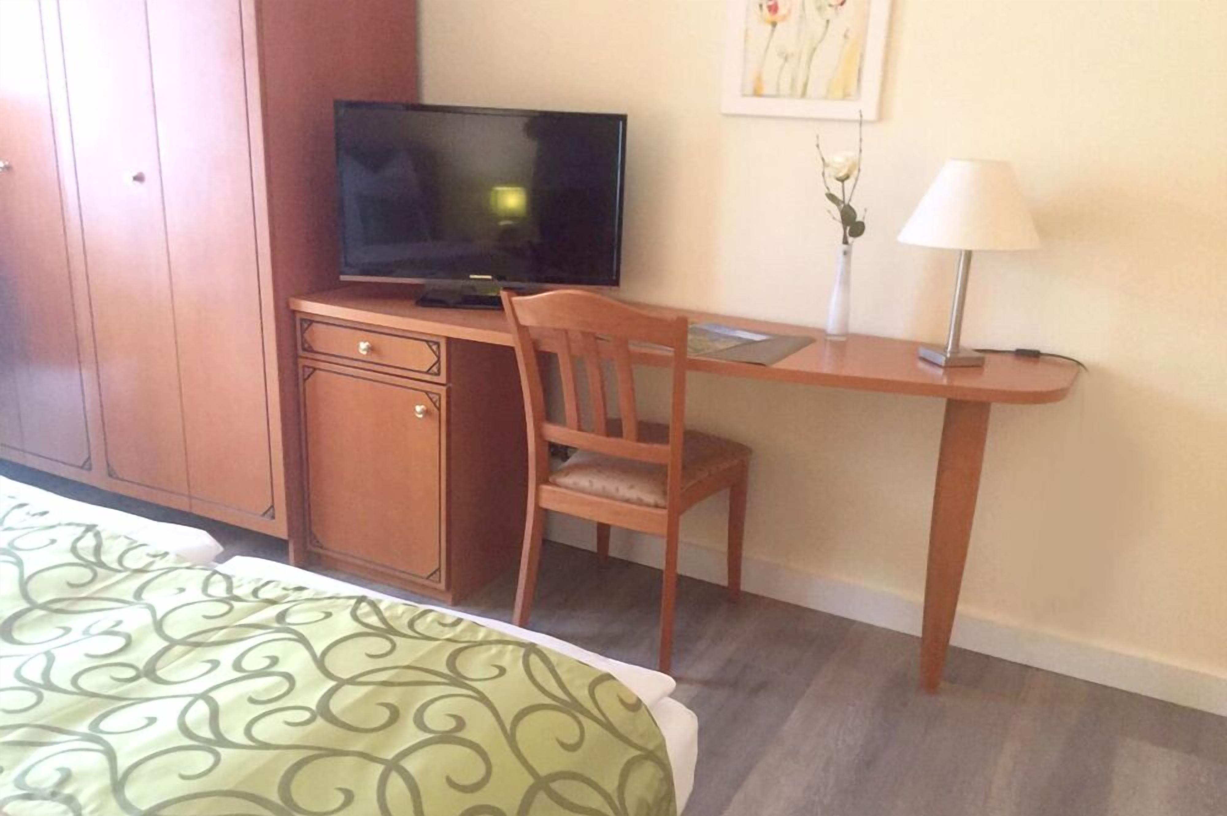 A hotel room with a bed, a desk with a television, a chair, a lamp, and a cabinet.