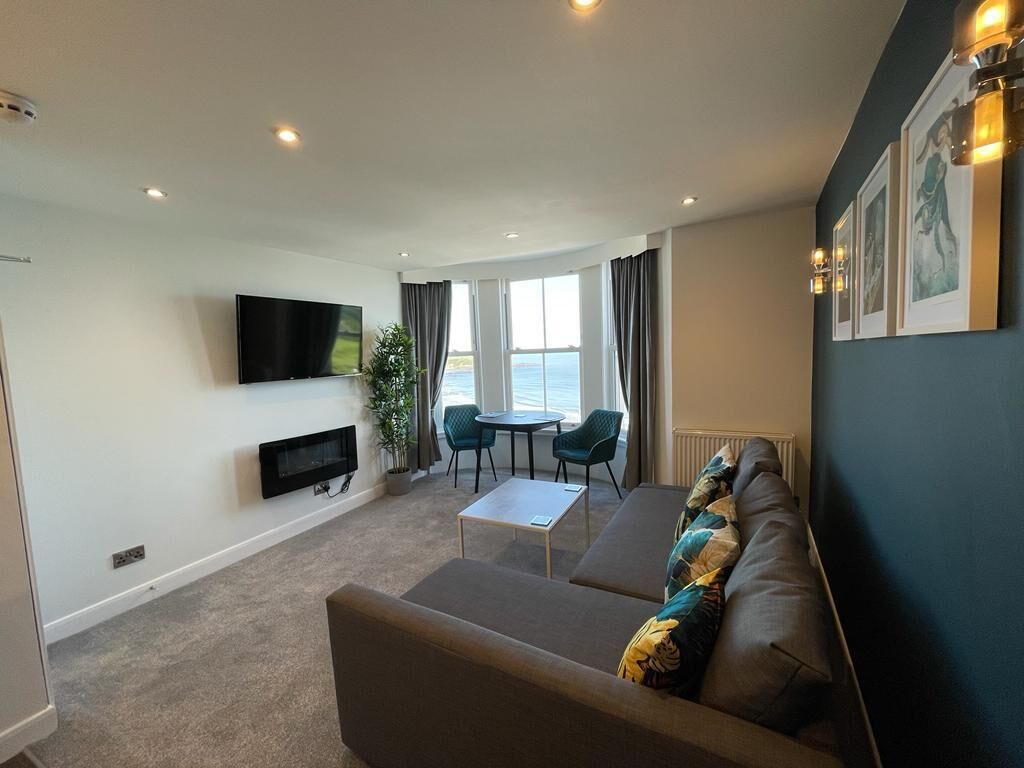 A modern living room with a sofa, a flat-screen TV, a small table, and a view of the sea through large windows.