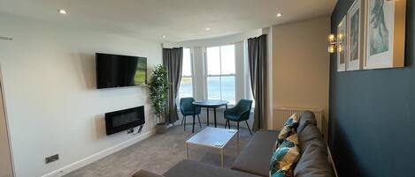 A modern living room with a sofa, a flat-screen TV, a small table, and a view of the sea through large windows.