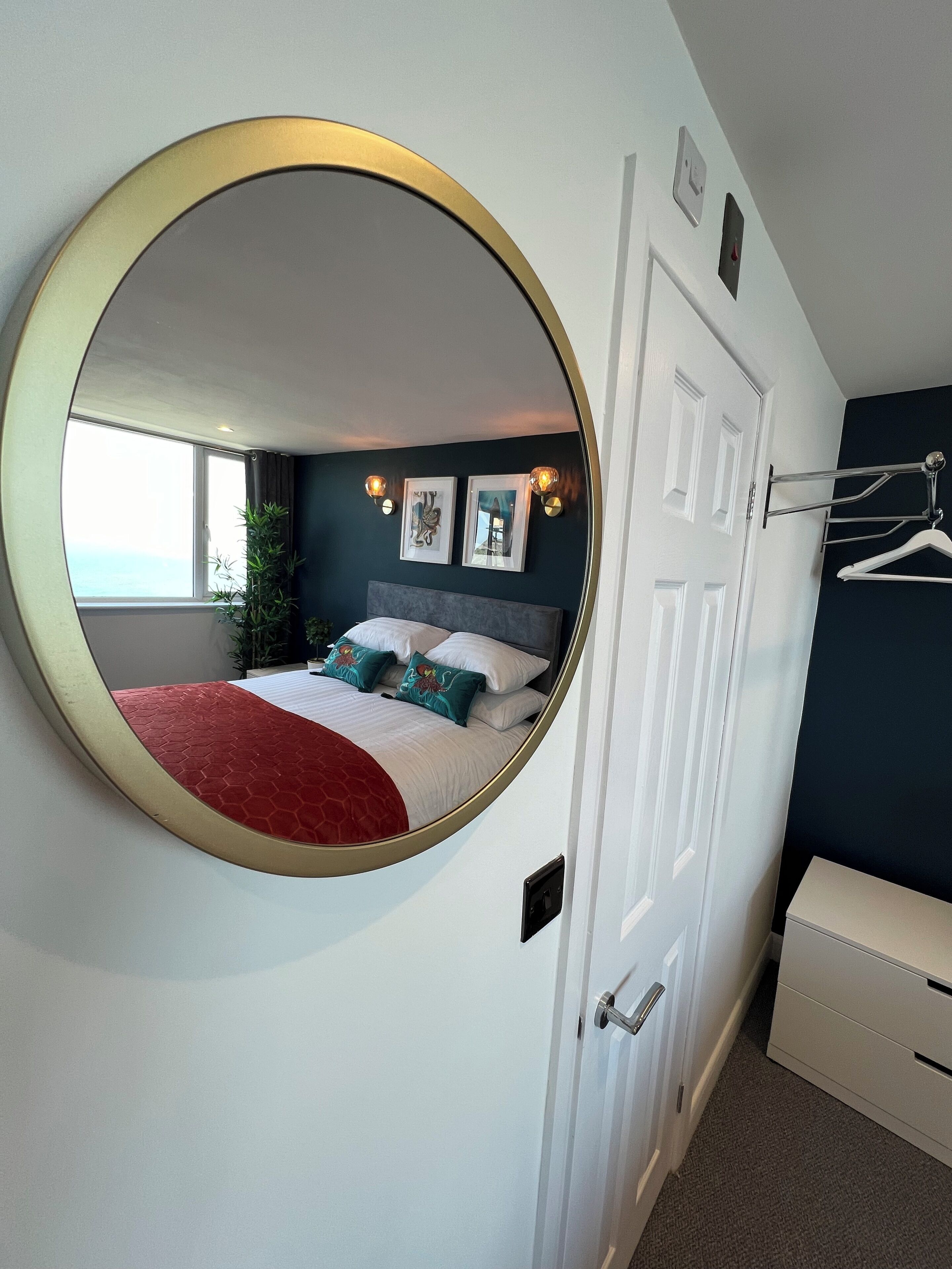A bedroom with a round mirror, a bed with a red blanket, a dresser, and a door with a coat hanger.