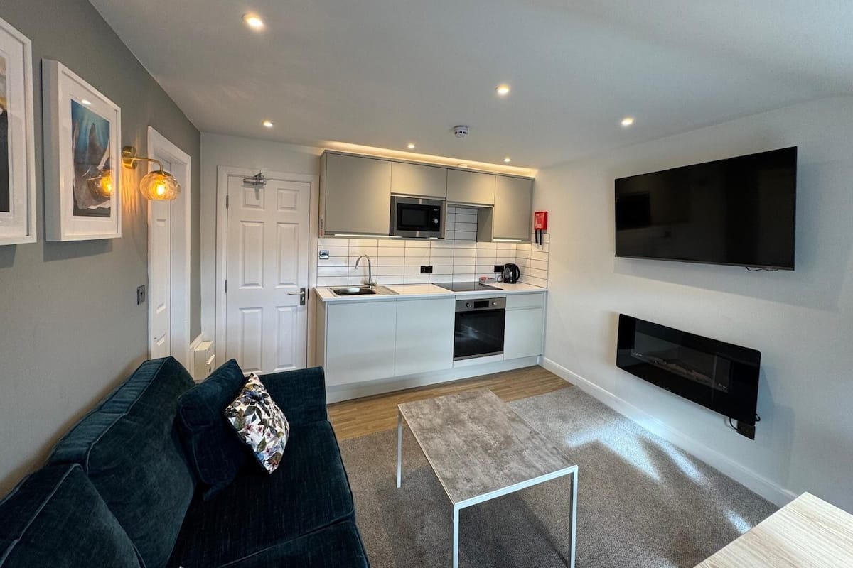 A modern kitchen with white cabinets, a microwave, and a sink. There is a dark green sofa with a floral pillow, a small white table, and a flat-screen TV mounted on the wall.