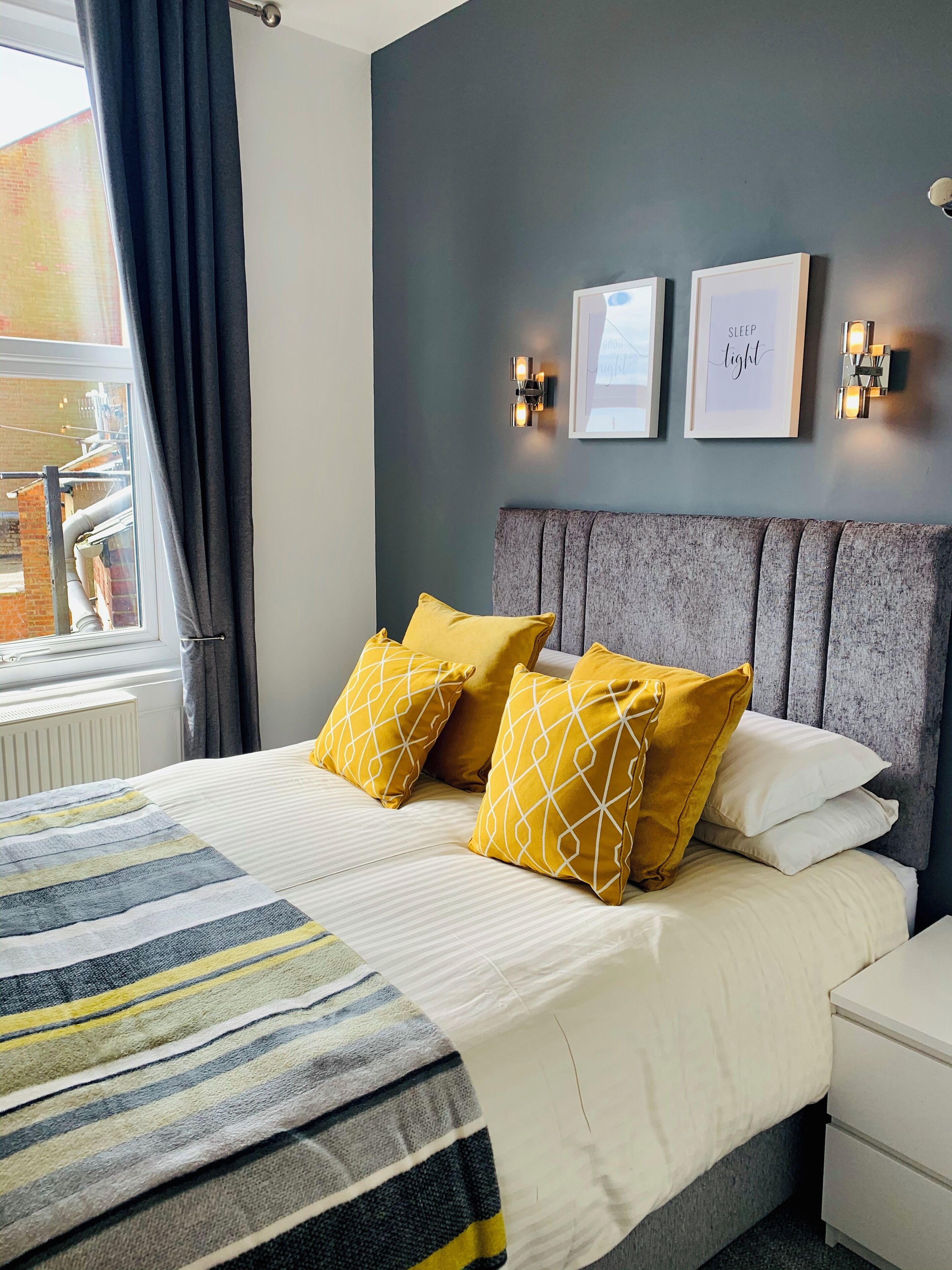 A bedroom with a bed, two pillows, a bedside table, a nightstand, and a window with curtains.