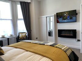A hotel room with a bed, a TV mounted on the wall, a fireplace, a window with curtains, and a small table with a bowl.