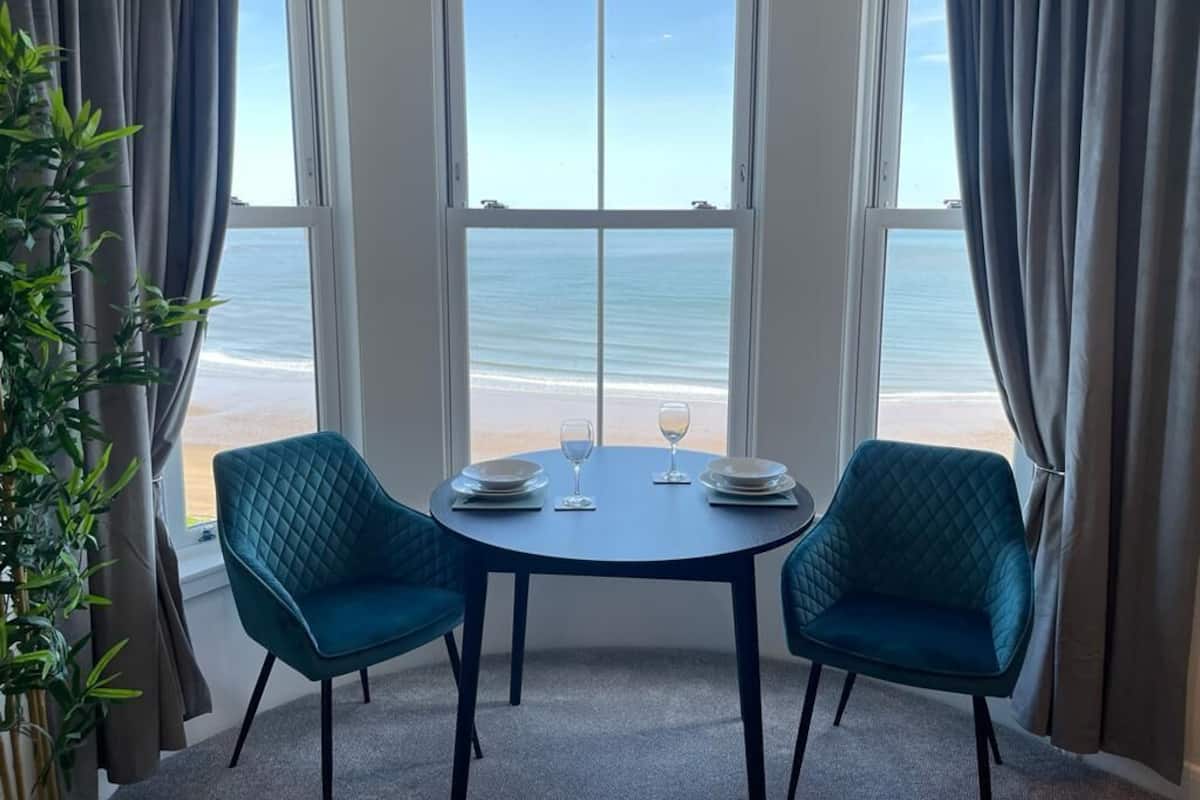A corner room with a large window overlooking the beach, two teal chairs, a round table, and a potted plant.