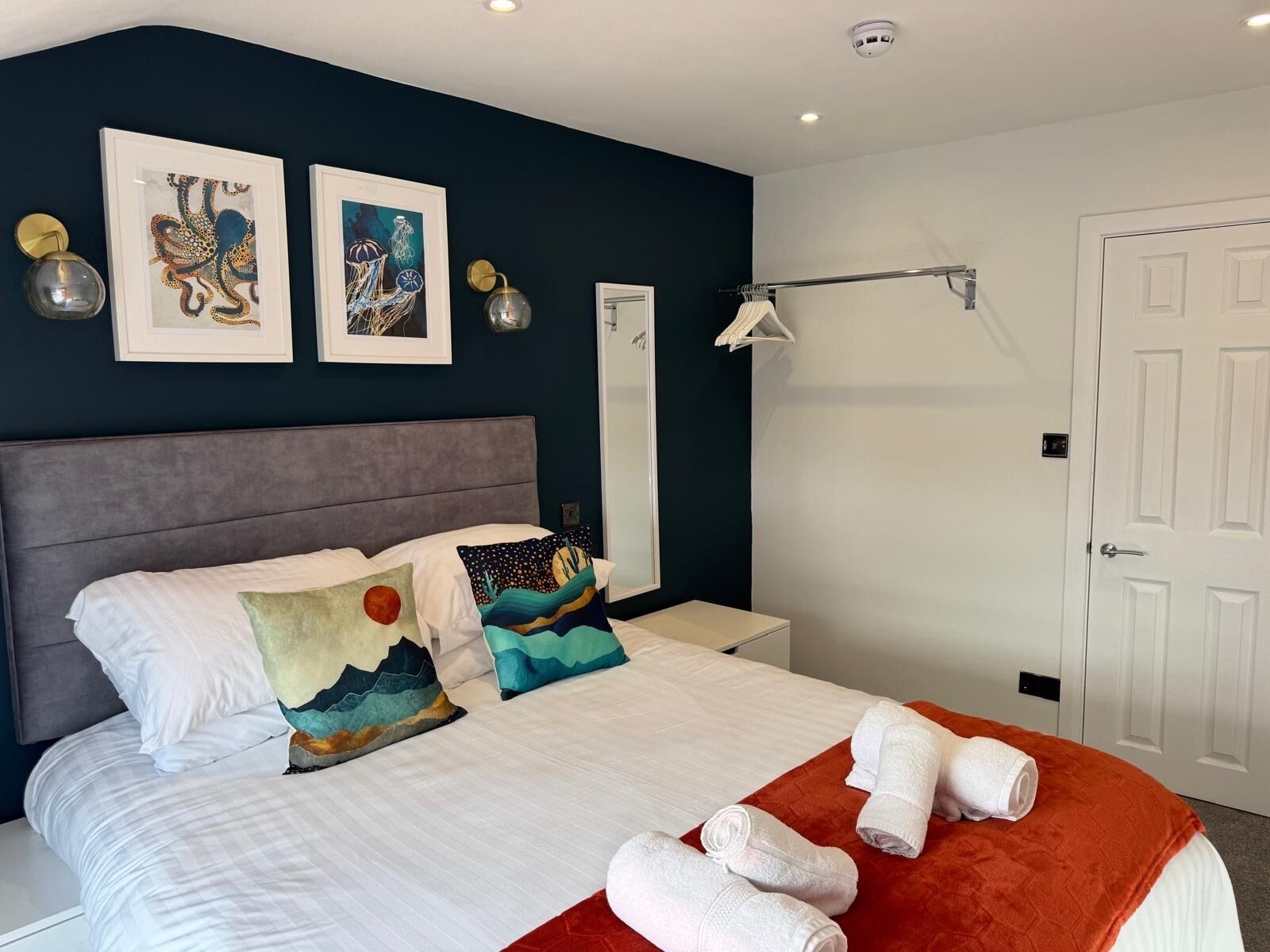 A neatly made bed with white linens, a grey headboard, and decorative pillows. There are two wall-mounted light fixtures and a white door in the background.