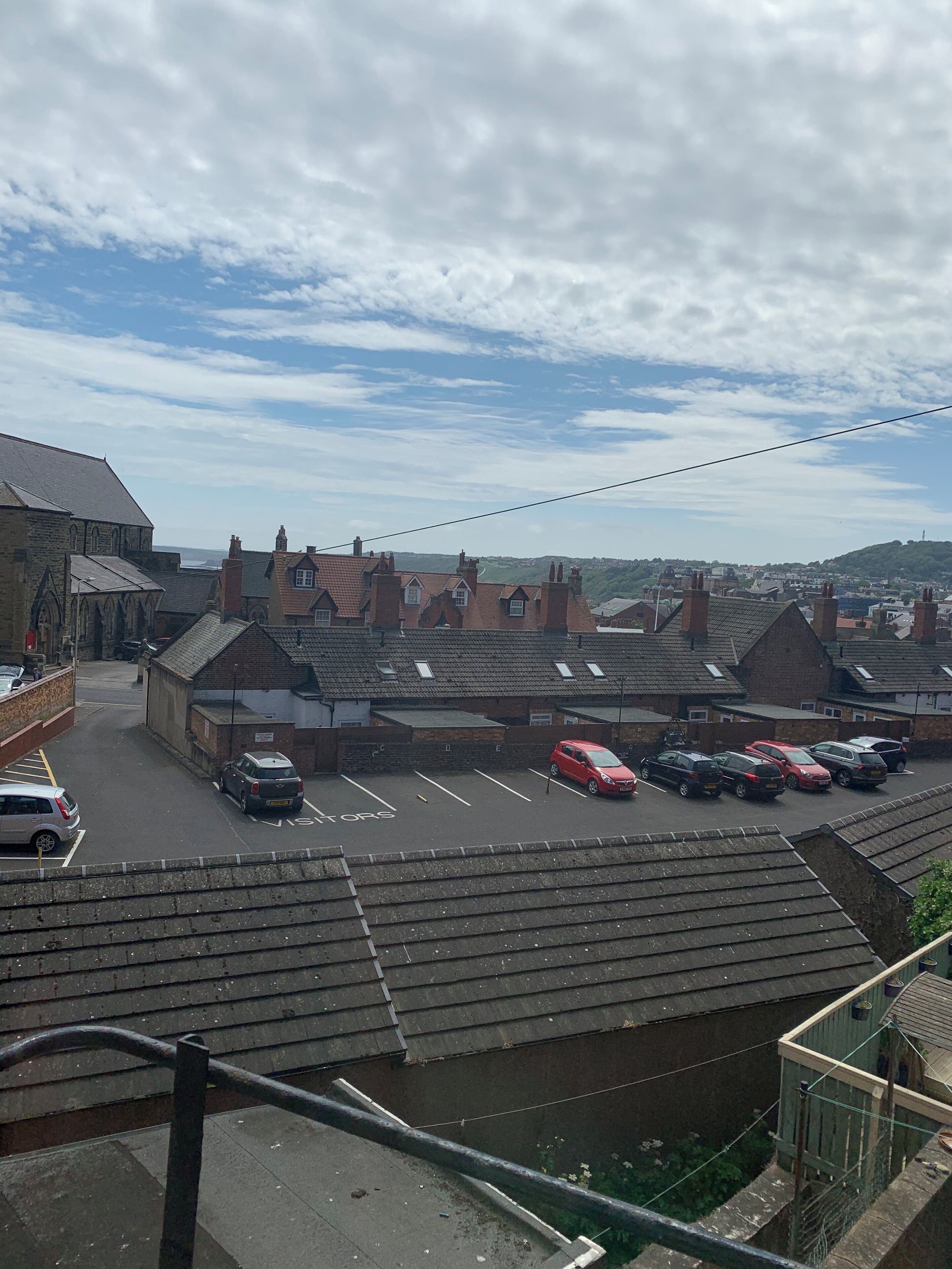 A parking lot with several cars, a building with a steep roof, and a view of the sea in the background.