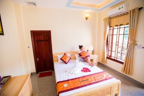 A hotel room with a bed, a wooden nightstand, a red doormat, a window with curtains, and an air conditioning unit.