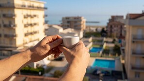 A person holding a white cup, overlooking a residential area with multiple buildings and a swimming pool.