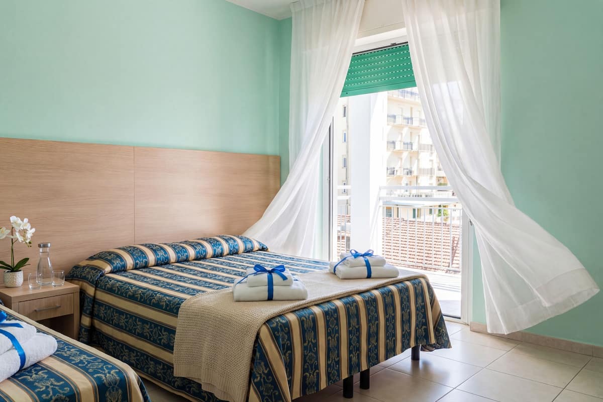 A hotel room with two beds, a nightstand, a vase with flowers, and a view of the city through sheer curtains.