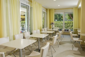 A dining area with tables and chairs, large windows with curtains, and a view of greenery outside.