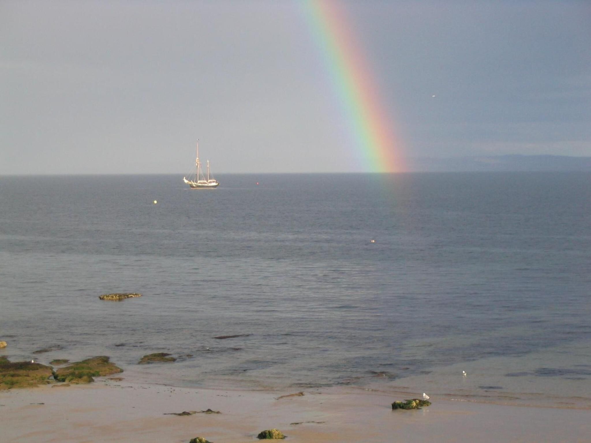 A beach with a sailboat in the distance and a rainbow in the sky.