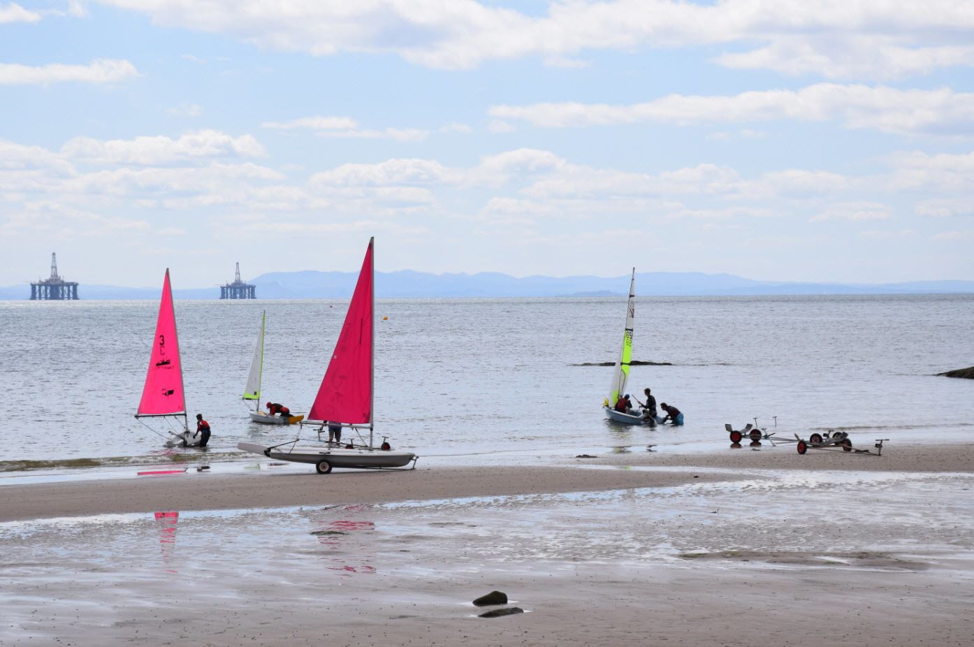 A beach with sailboats and a distant offshore platform.