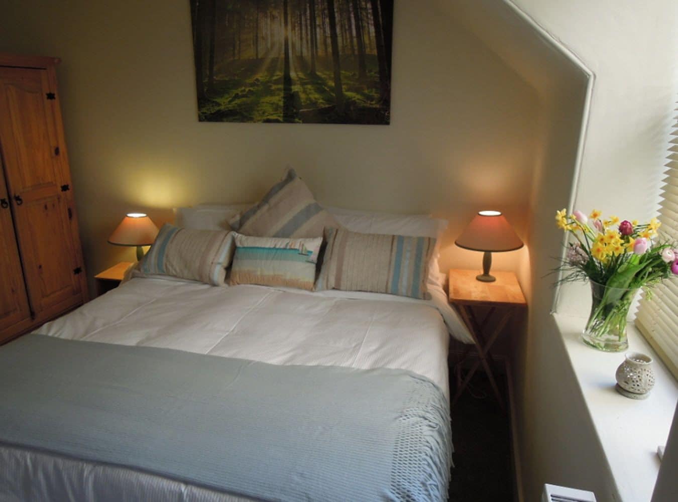 A neatly made bed with pillows, a wooden wardrobe, a painting of a forest, two bedside lamps, and a vase with flowers.