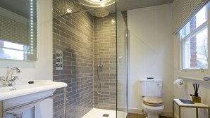 A modern bathroom with a glass-enclosed shower, a white sink with a mirror, a toilet with a wooden seat, and a small table with toiletries. - The Northgate (Bury St Edmunds)