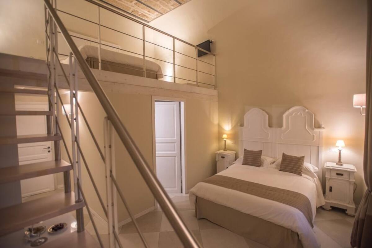 Un dormitorio con una cama doble, una mesita de noche, una lámpara de noche, una escalera y una puerta que da a otra habitación.
