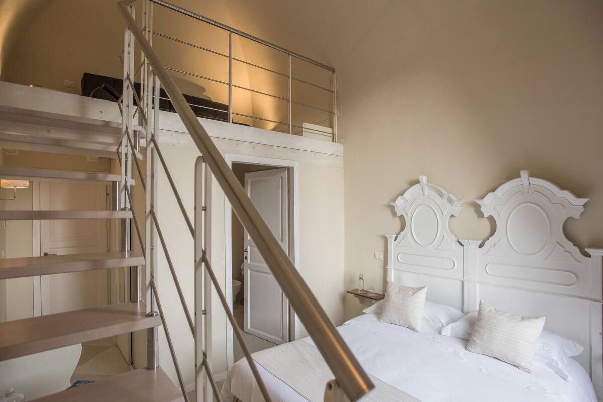 Un dormitorio con una cama blanca, una escalera de madera y un cabecero blanco.